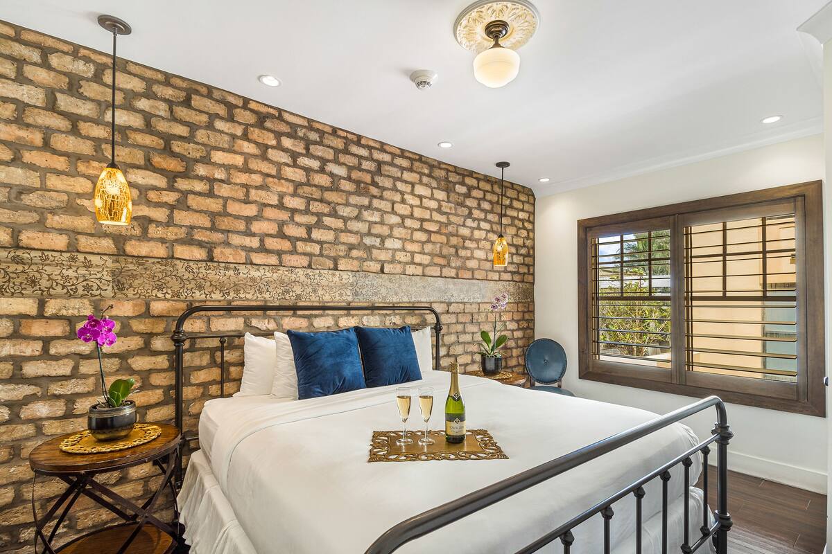 A neatly made bed with a bottle and two glasses on a tray, a brick wall, a ceiling fan, and a window with blinds.