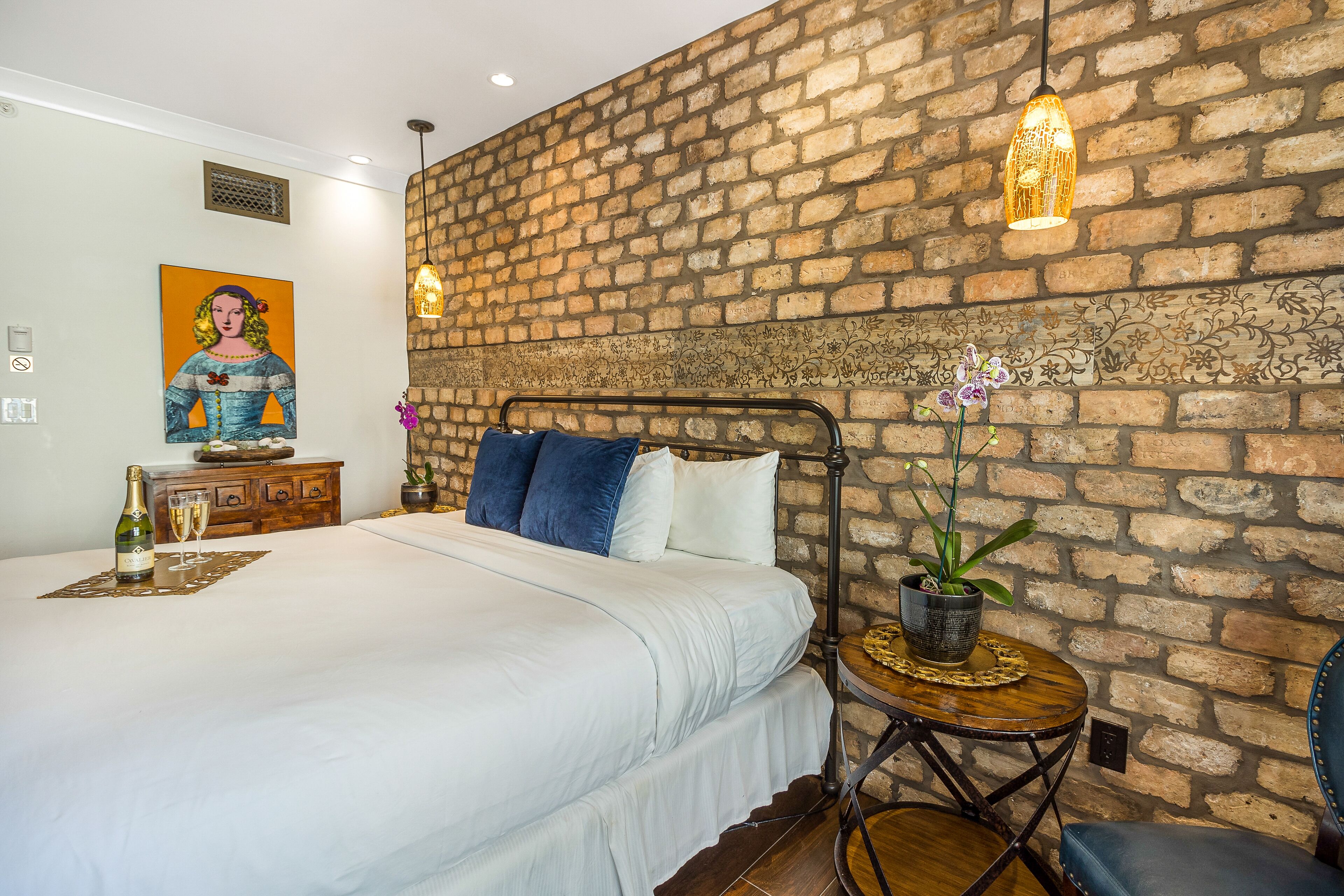 A bedroom with a brick wall, a bed with white linens, a nightstand with a bottle and glasses, and a small table with a plant.