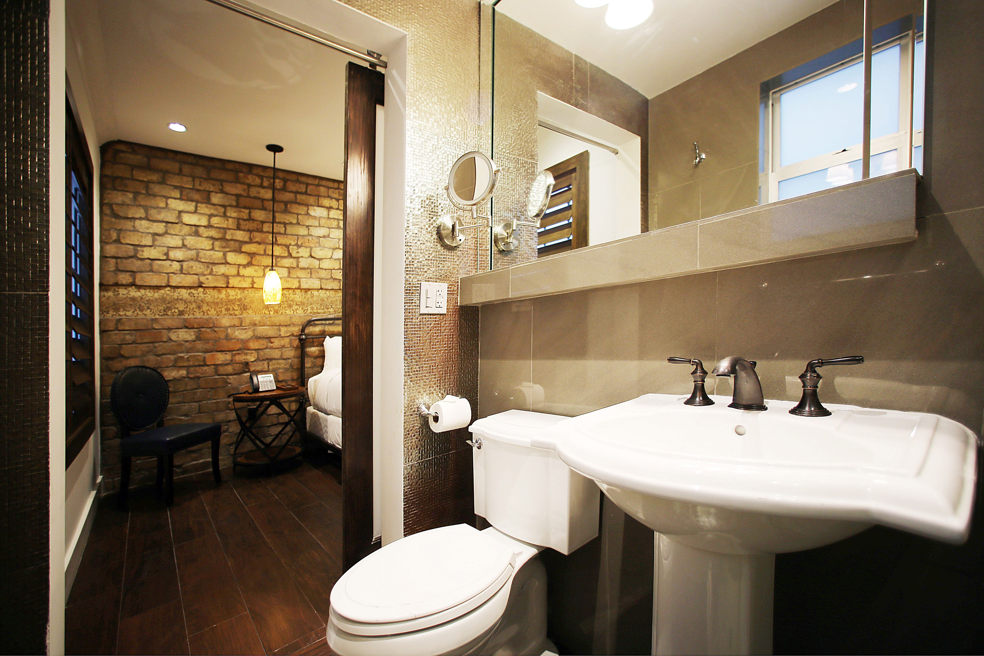 Une salle de bain avec des toilettes blanches, un grand lavabo et un miroir au-dessus. On aperçoit un mur de briques et une fenêtre à l’arrière-plan.