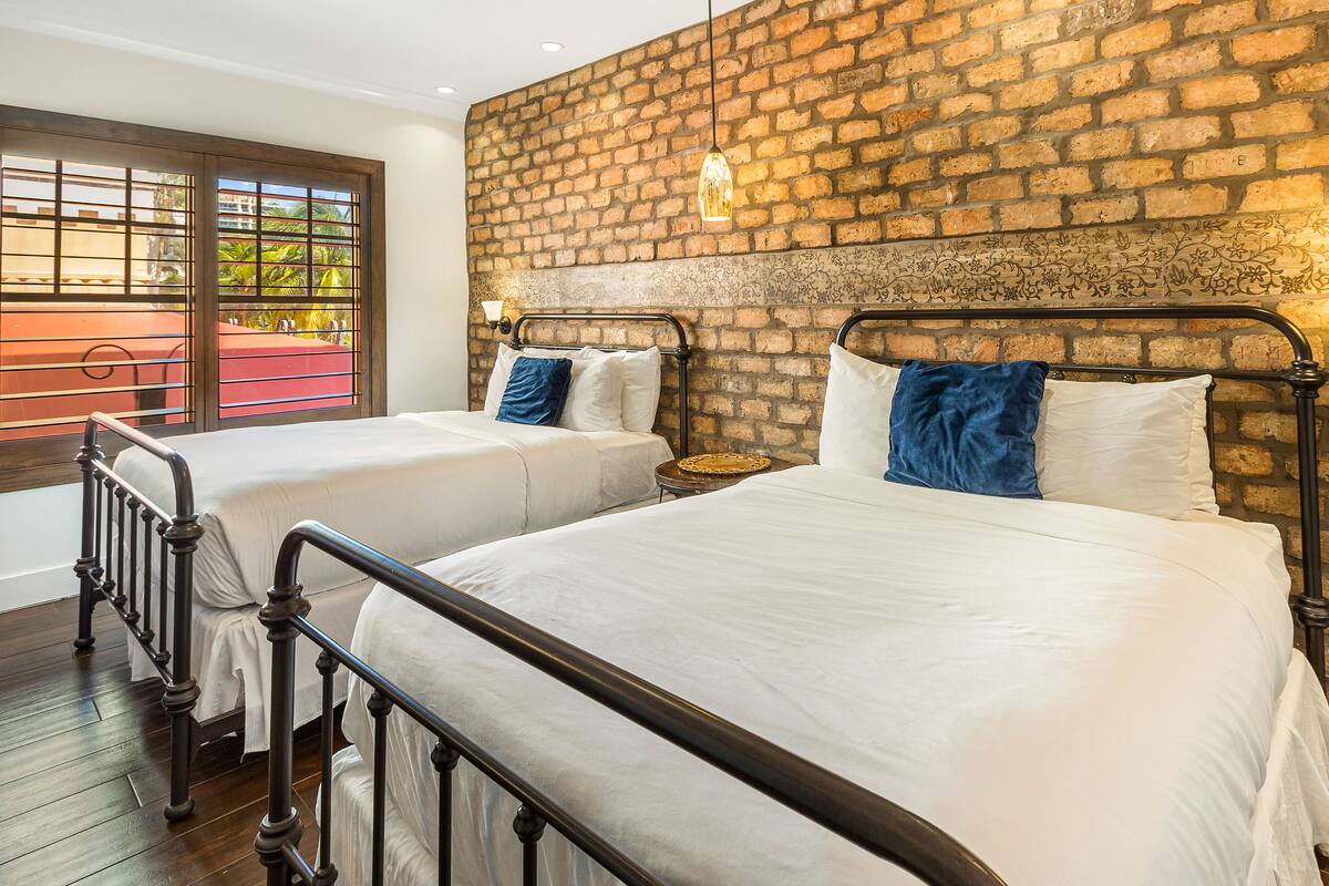 A room with two beds, a brick wall, and a window with a view of greenery.