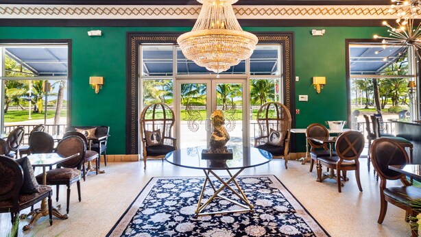 A hotel lobby with a large chandelier, green walls, and a view of palm trees through the glass doors.