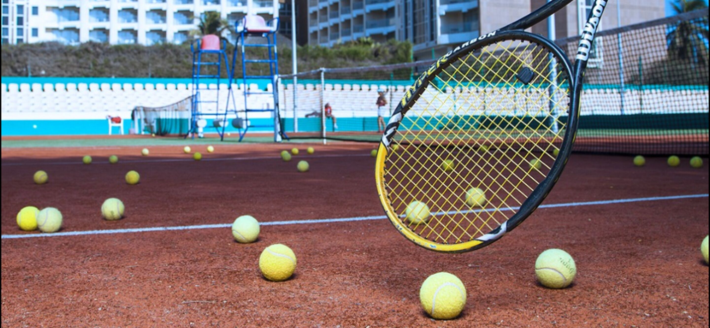 A tennis court with a tennis racket and multiple tennis balls.