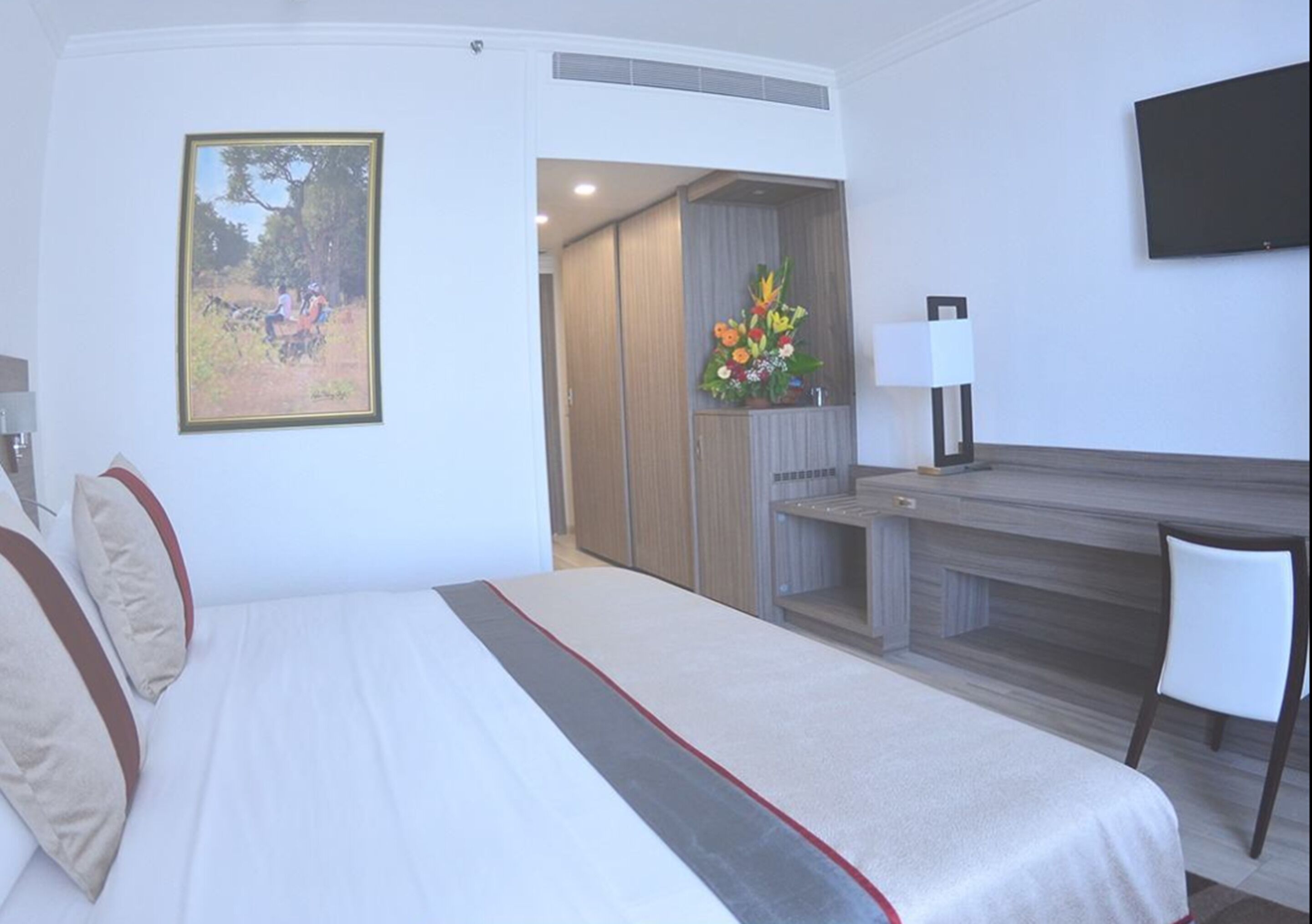 A hotel room with a bed, a desk, a chair, a TV, and a flower arrangement.