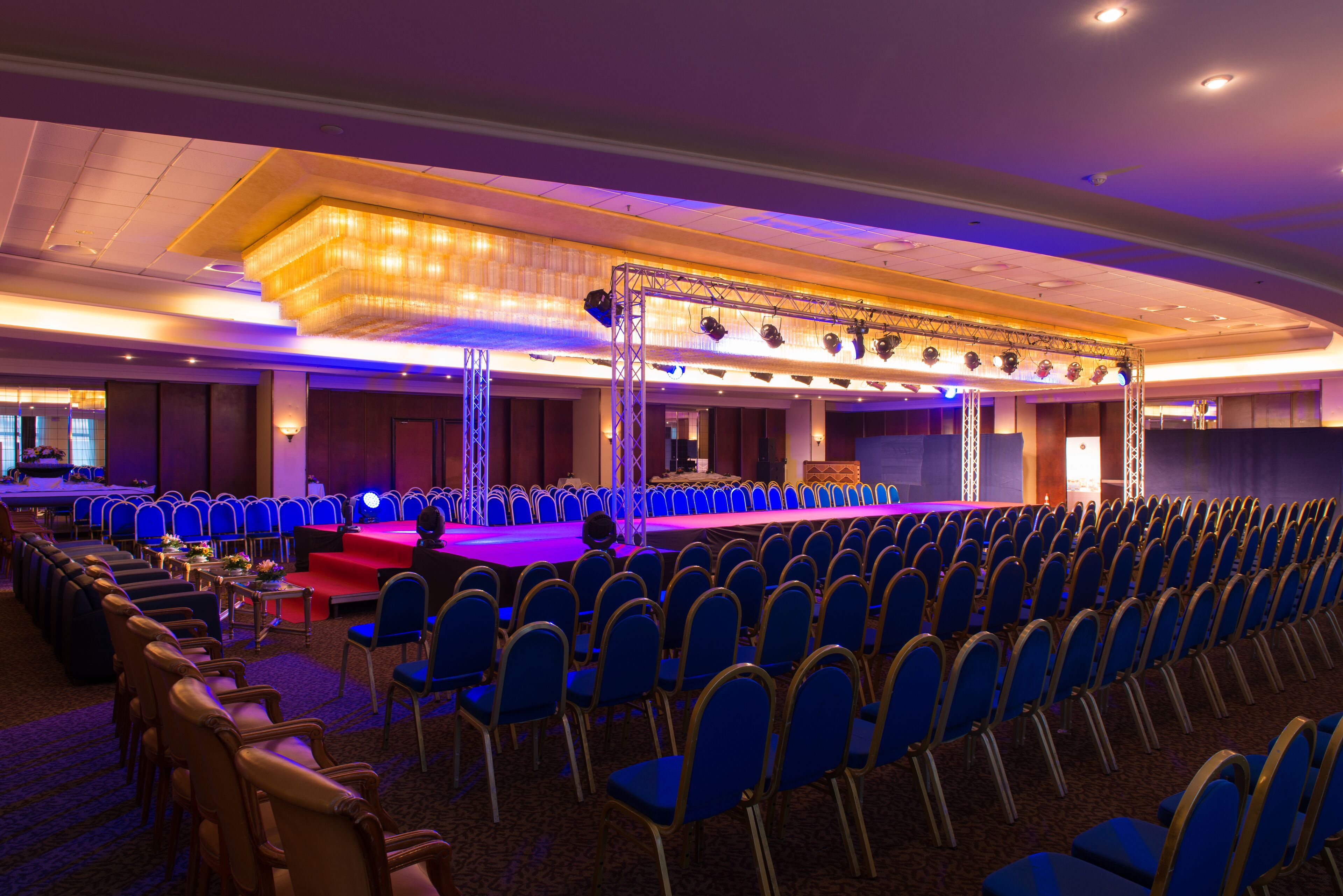 Un amplio salón de conferencias con filas de sillas azules, un escenario con fondo rojo y una lámpara de araña.