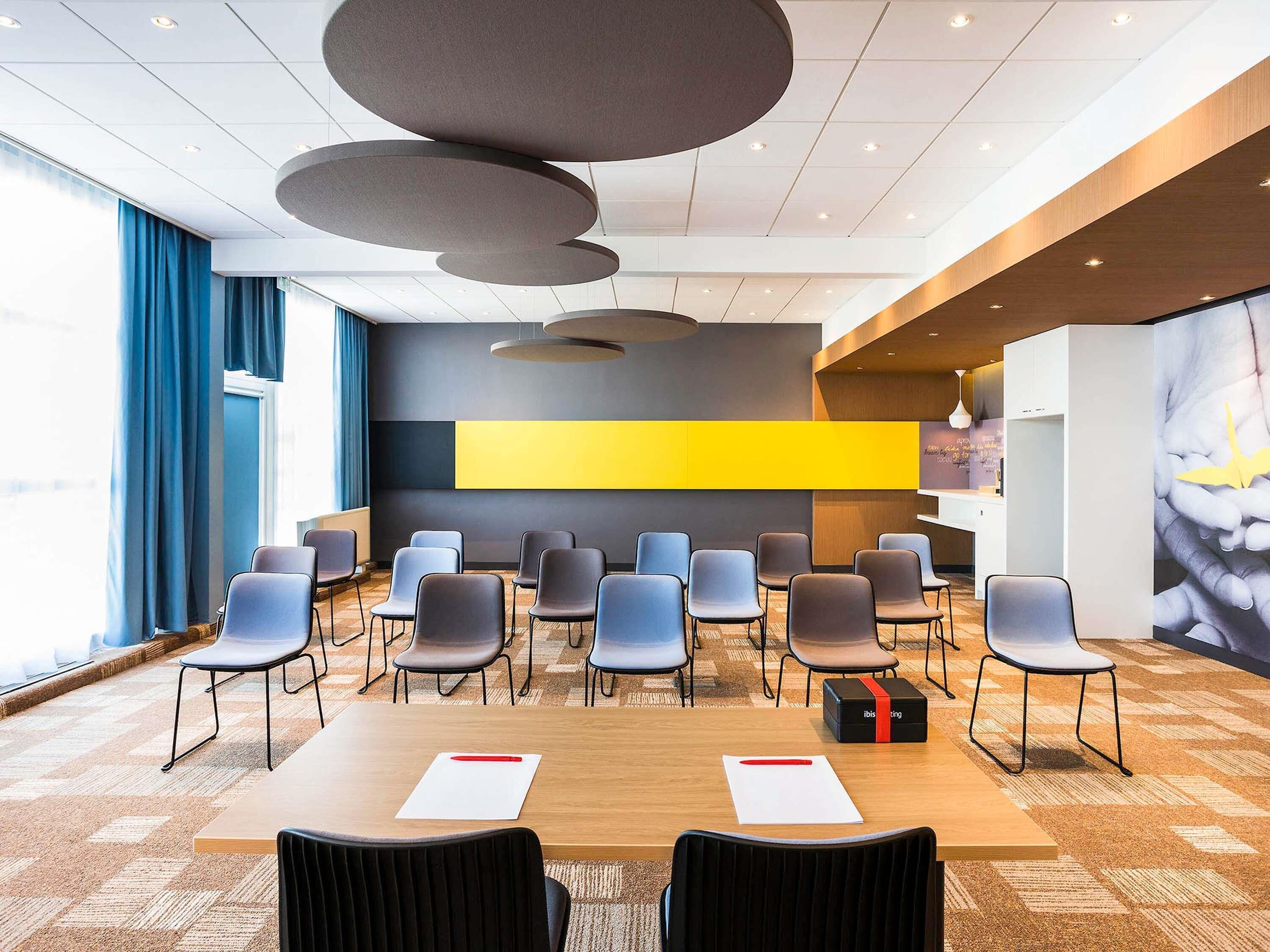 A conference room with a long table, multiple chairs, a large yellow accent wall, and a modern ceiling design.
