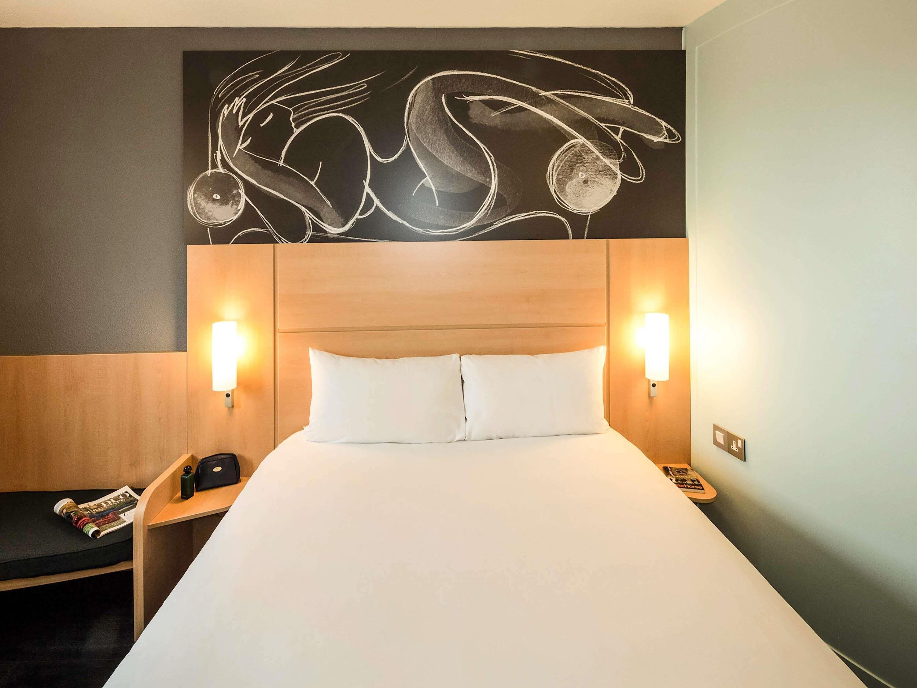 Une chambre d’hôtel avec un grand lit, deux lampes de chevet, une tête de lit en bois et une œuvre d’art décorative accrochée au mur au-dessus du lit.
