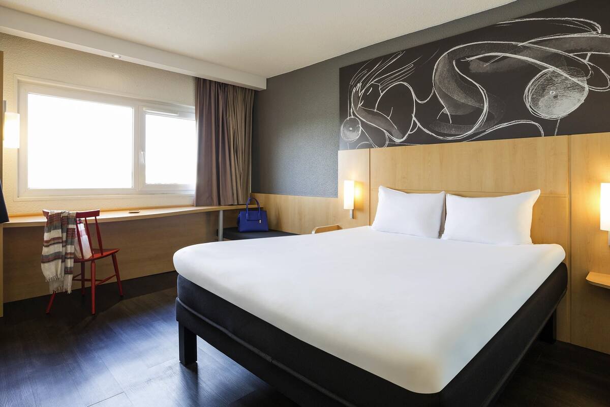 Ein Hotelzimmer mit einem großen Bett, einem Schreibtisch mit Stuhl, einem Fenster mit Vorhängen und einer Wand mit einer Kreidezeichnung.