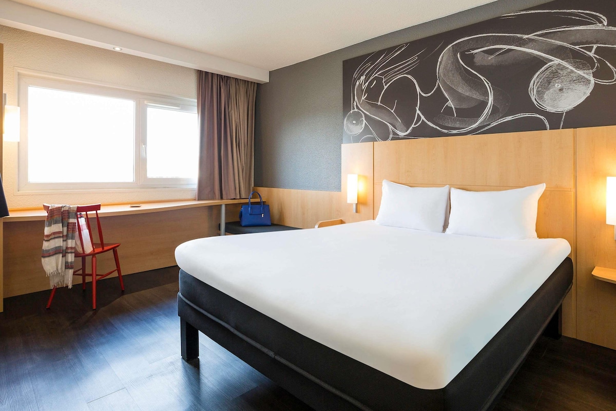 A hotel room with a large bed, a desk with a chair, a window with curtains, and a wall with a chalkboard drawing.