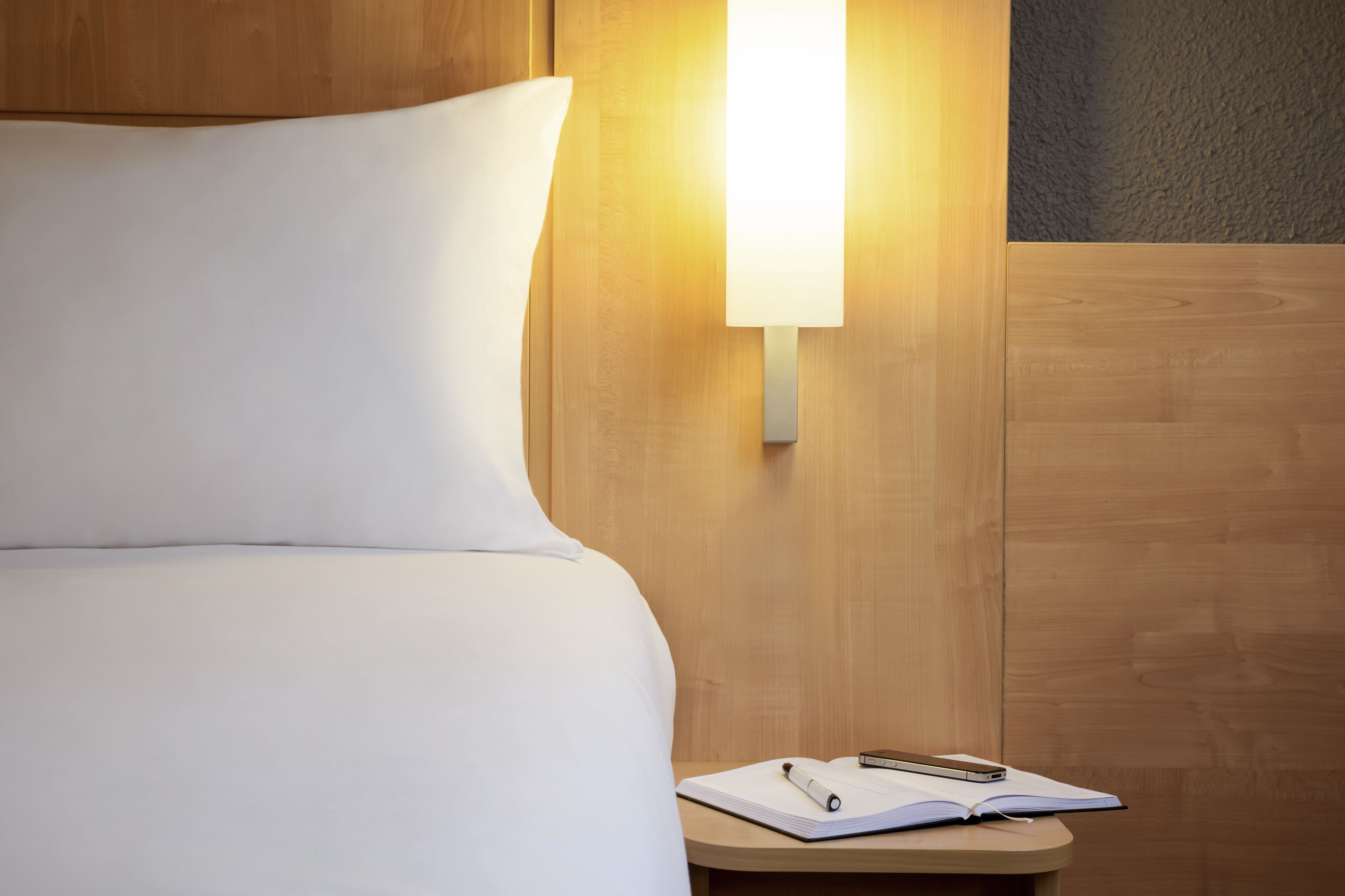 A neatly made bed with white bedding, a bedside table with a notebook, a pen, and a smartphone.