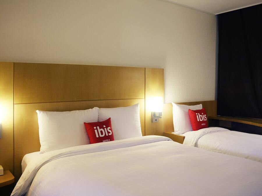 Een hotelkamer met twee bedden, elk voorzien van witte beddengoed en rode kussens met de tekst 'ibis'. Aan beide zijden van de bedden bevinden zich wandlampen, en boven de bedden hangt een flatscreen-televisie.