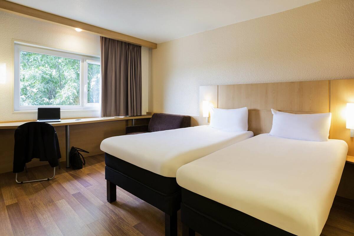 Ein Hotelzimmer mit zwei Betten, einem Schreibtisch mit einem Laptop, einem Stuhl und einem Fenster mit Vorhängen.