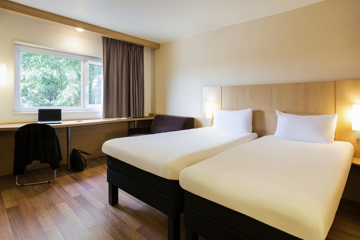 A hotel room with two beds, a desk with a laptop, a chair, and a window with curtains.