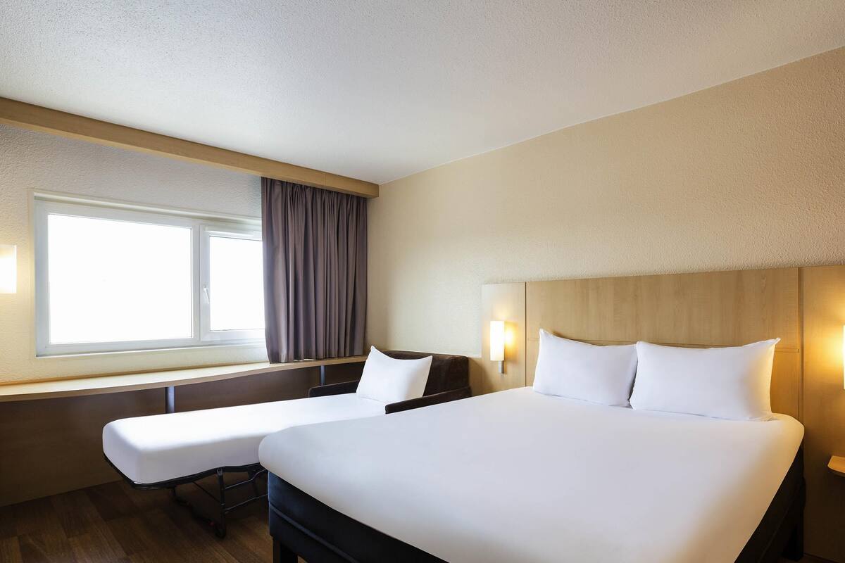 Ein Hotelzimmer mit einem großen Bett, einem Nachttisch, einem Sessel, einem Fenster mit Vorhängen und einer Wandleuchte.