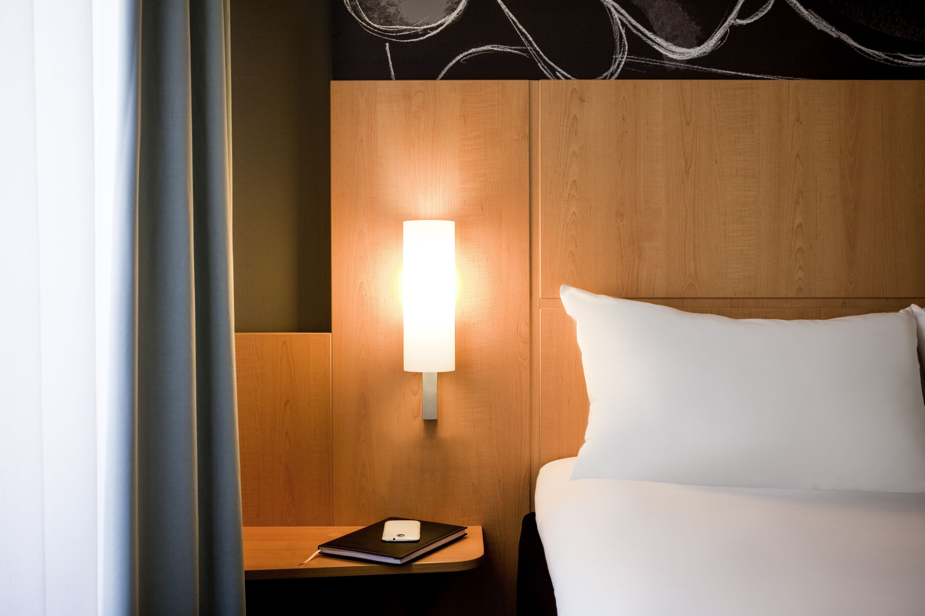 A hotel room with a wooden headboard, a bedside lamp, a white bed, and a nightstand with a notebook and a pen.