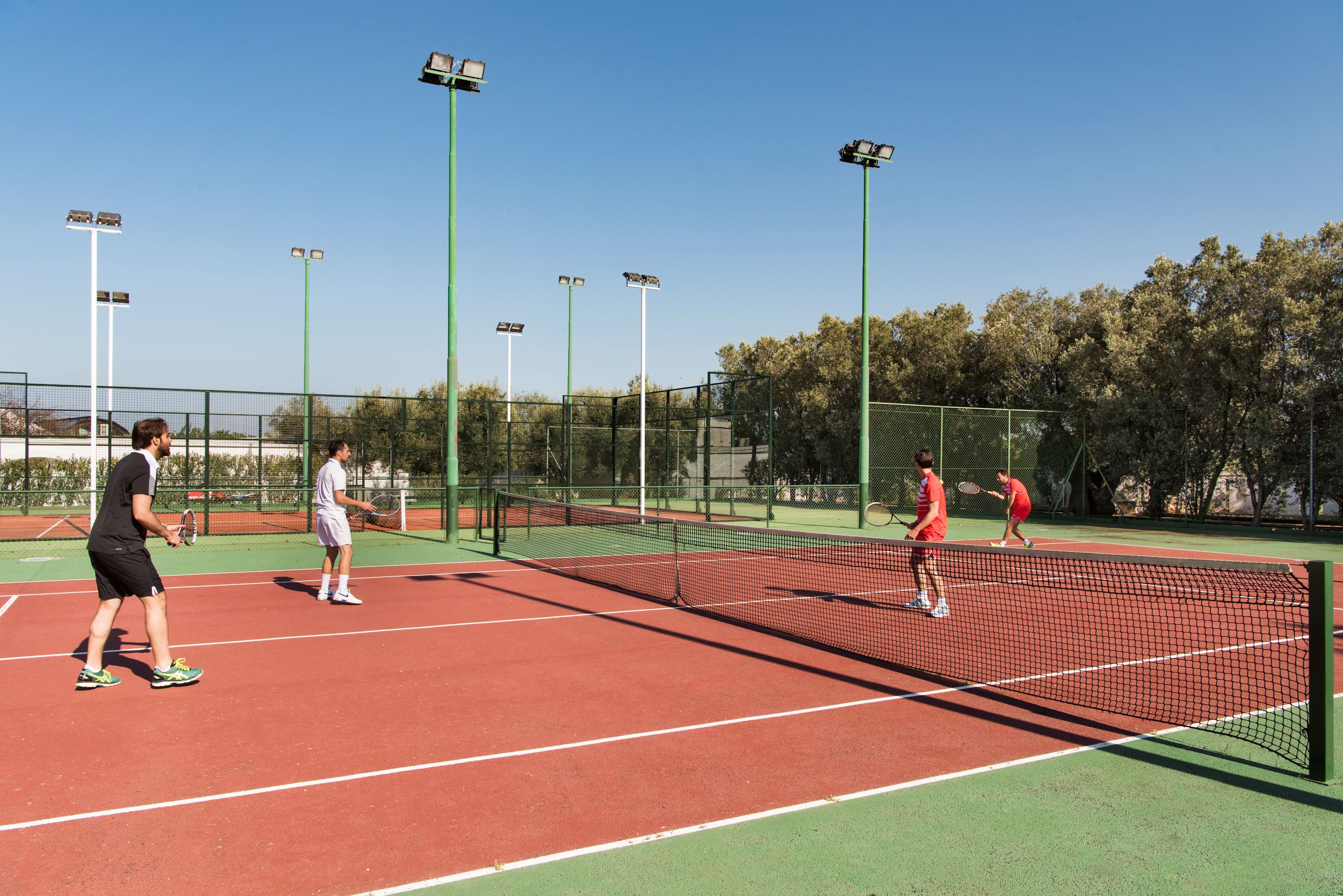 Ein Tennisplatz mit vier Spielern, grüner Umzäunung und hohen Flutlichtmasten.