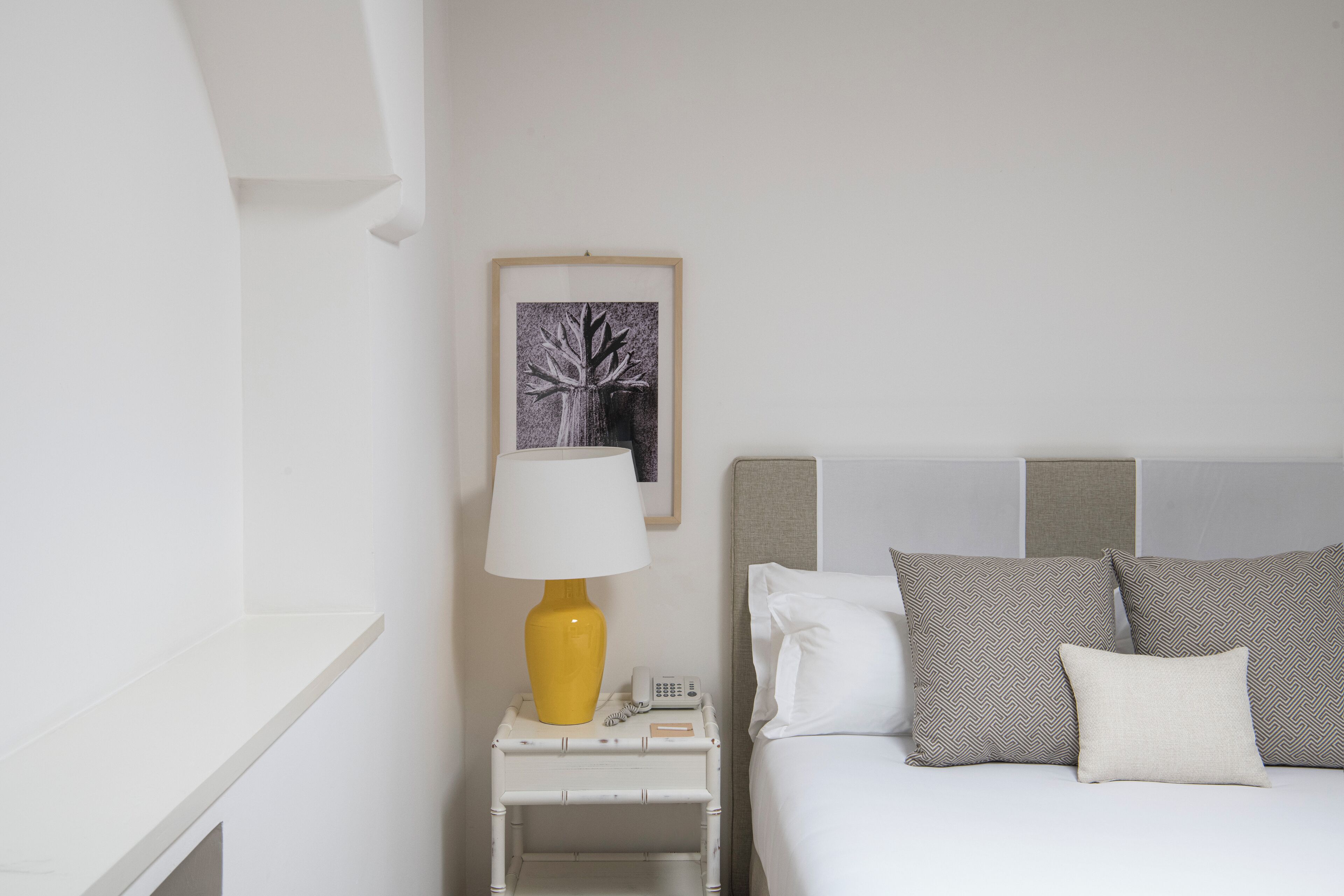 A neatly arranged hotel room with a bed, a bedside table with a lamp, a framed picture on the wall, and a bedside telephone.
