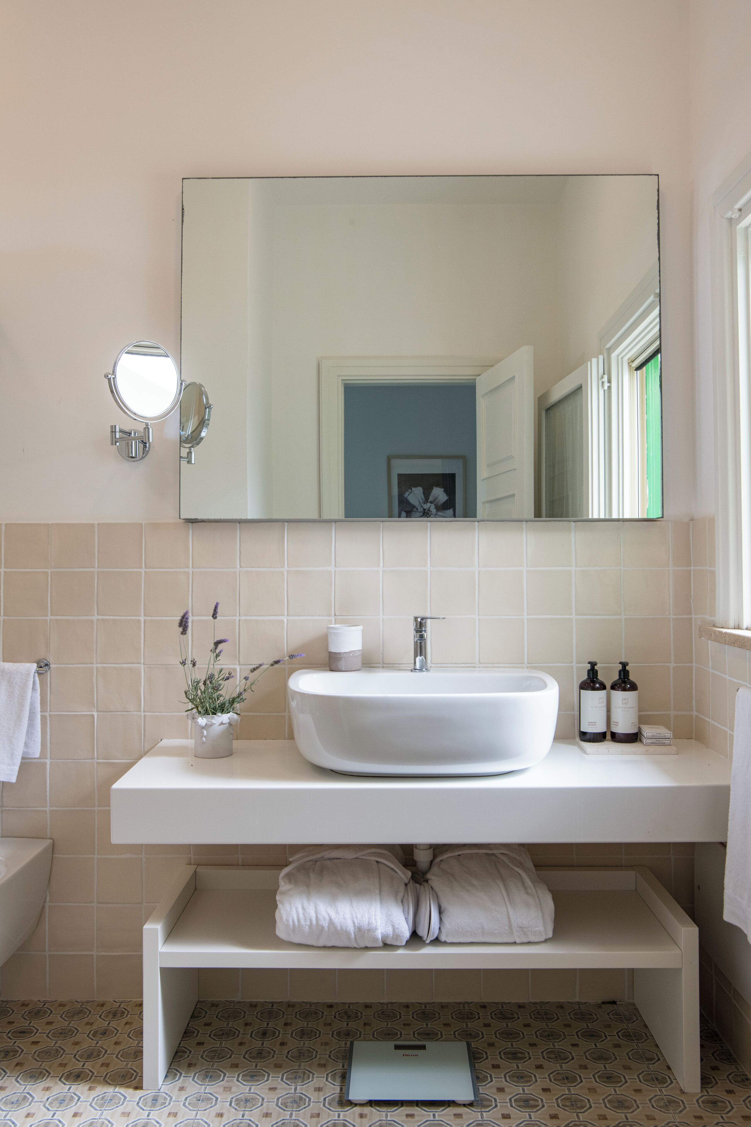 A bathroom with a white sink, mirror, and shelves. There is a towel rack, a small plant, and toiletries on the counter.
