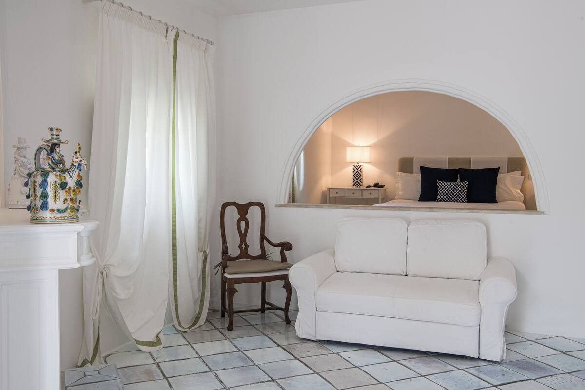 Une pièce avec un canapé blanc, une chaise en bois, un vase décoratif et une fenêtre ornée d’un rideau blanc.