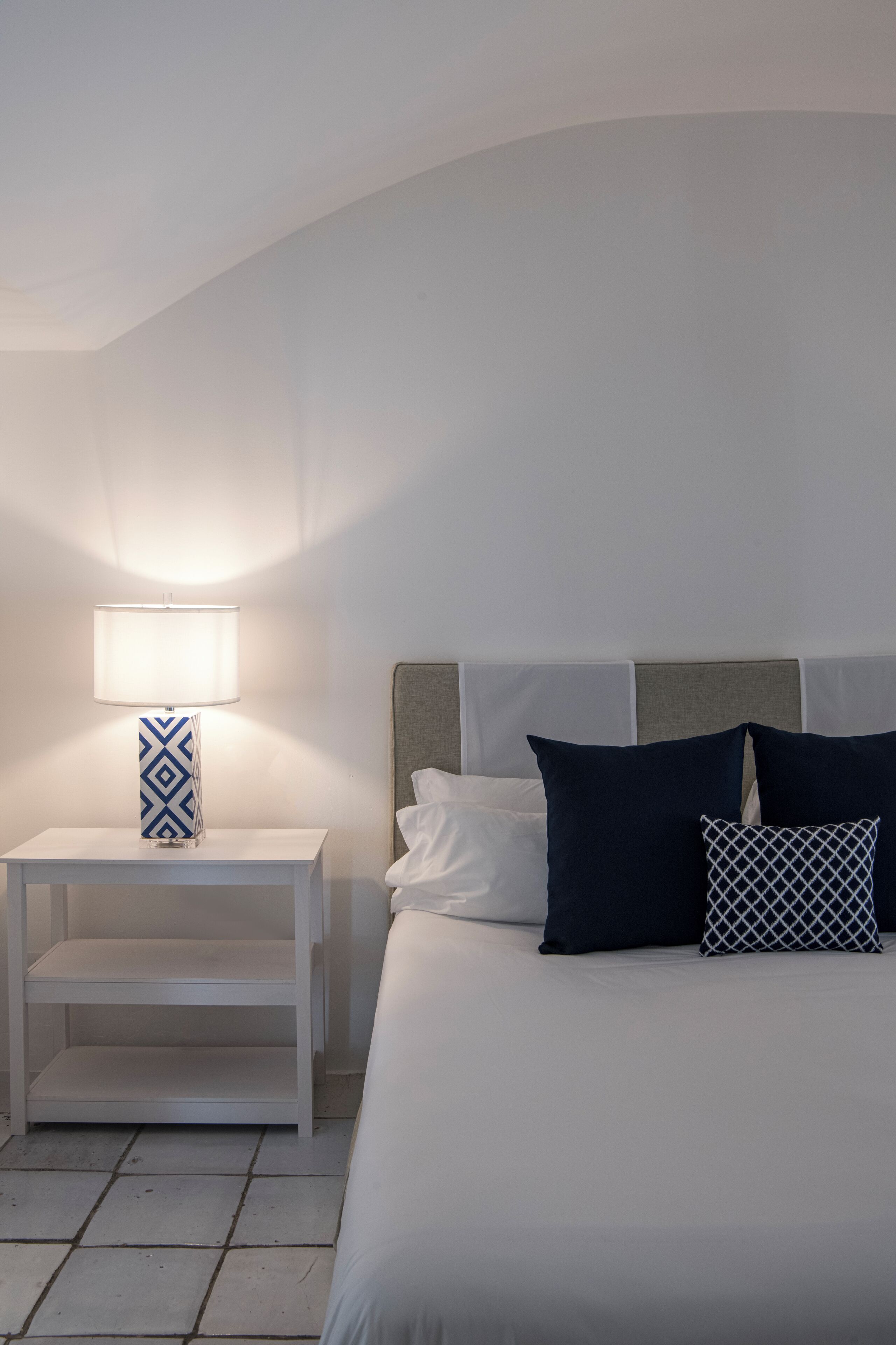 Un dormitorio ordenado con cama, mesita de noche blanca y lámpara con base de diseño geométrico.