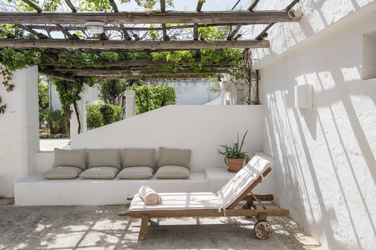 Un espace extérieur ensoleillé, aménagé avec des sièges, comprenant un fauteuil de jardin en bois avec des coussins, un banc blanc également doté de coussins, et une plante en pot.