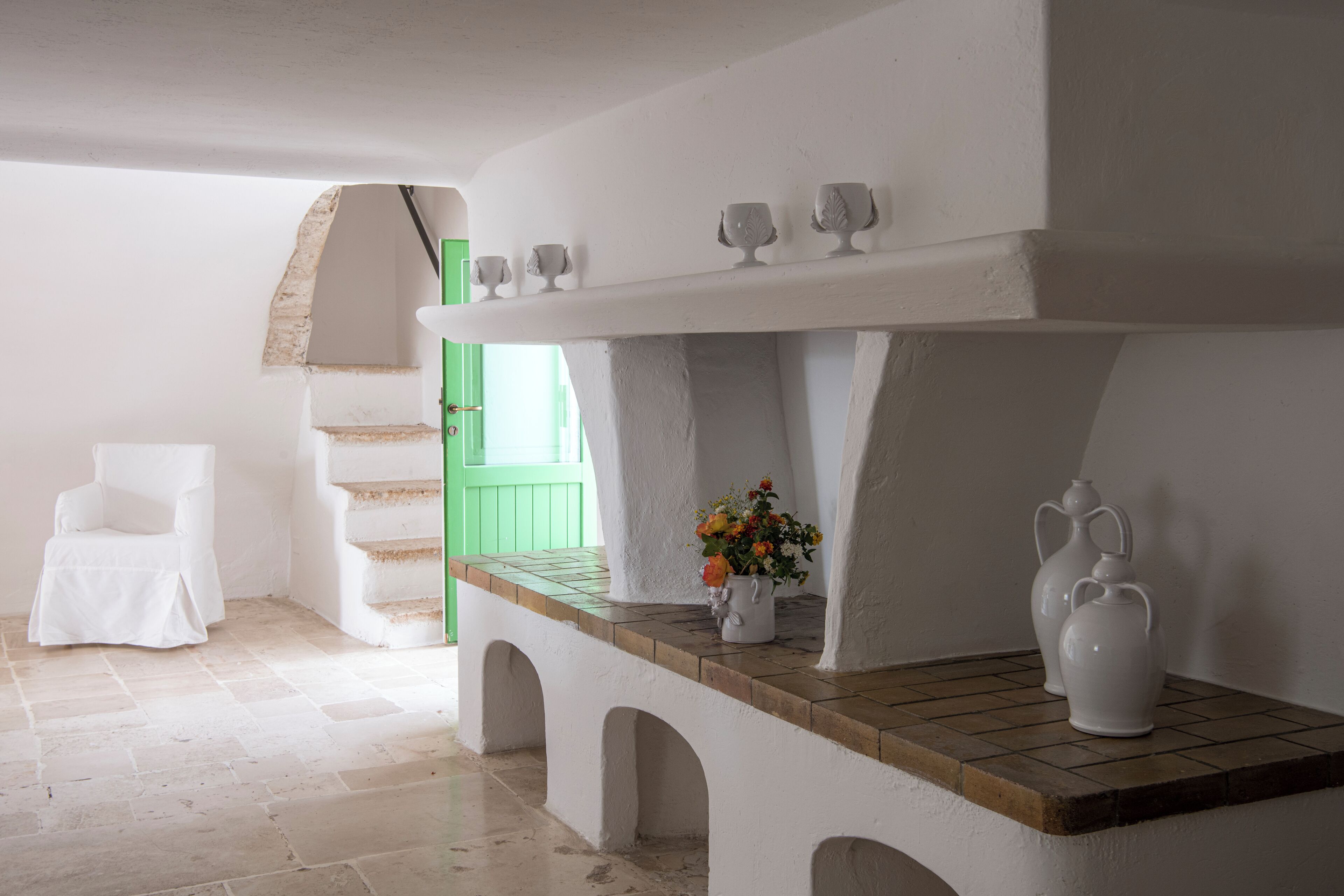 Una habitación con una silla blanca, una mesa de madera con un jarrón de flores y una jarra blanca.