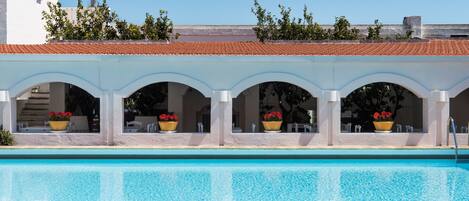 Uma piscina com deck ladrilhado, área de estar coberta com arcos e plantas em vasos.