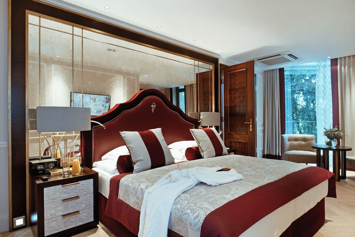 A spacious bedroom with a large bed, a nightstand with a lamp, and a view of the outdoors through a window.