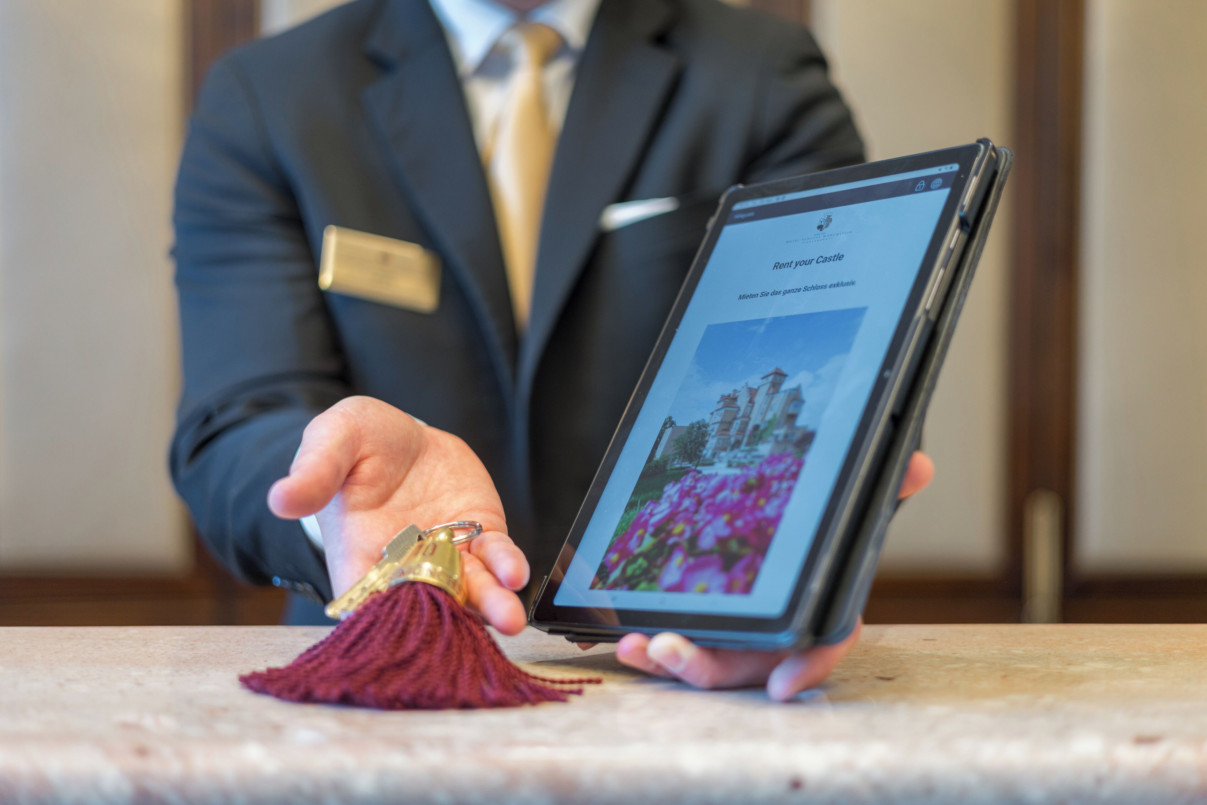 Eine Person hält ein Tablet, auf dem ein Bild eines Schlosses zu sehen ist. Ein Schlüsselanhänger und ein Namensschild sind sichtbar.