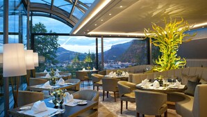 Un restaurante con mesas dispuestas para comer, un jarrón grande con flores amarillas y vistas a montañas y un pueblo.