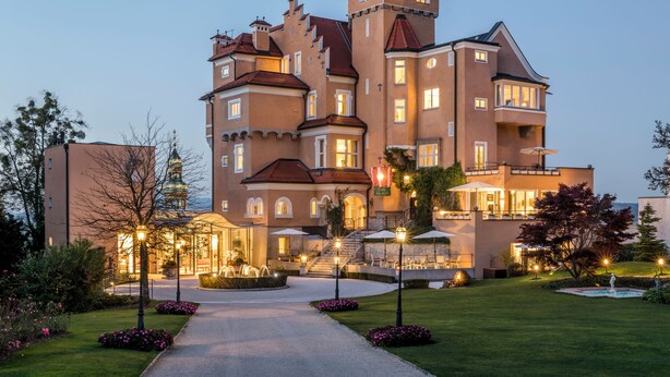 Un hotel grande de varios pisos con un patio central, mobiliario exterior e ingreso bien iluminado.