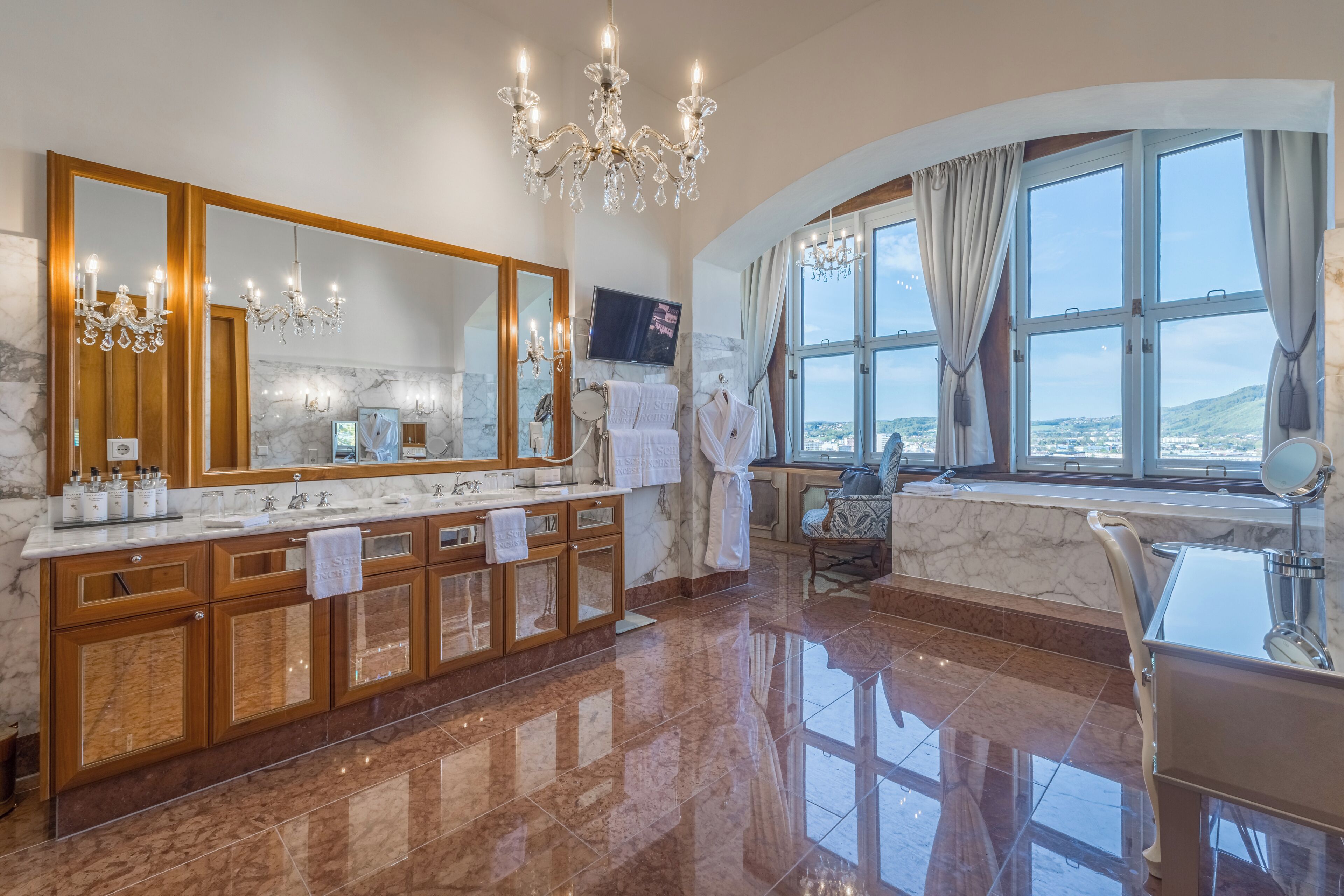 A luxurious bathroom with a marble vanity, a large mirror, a chandelier, and a view through a window.