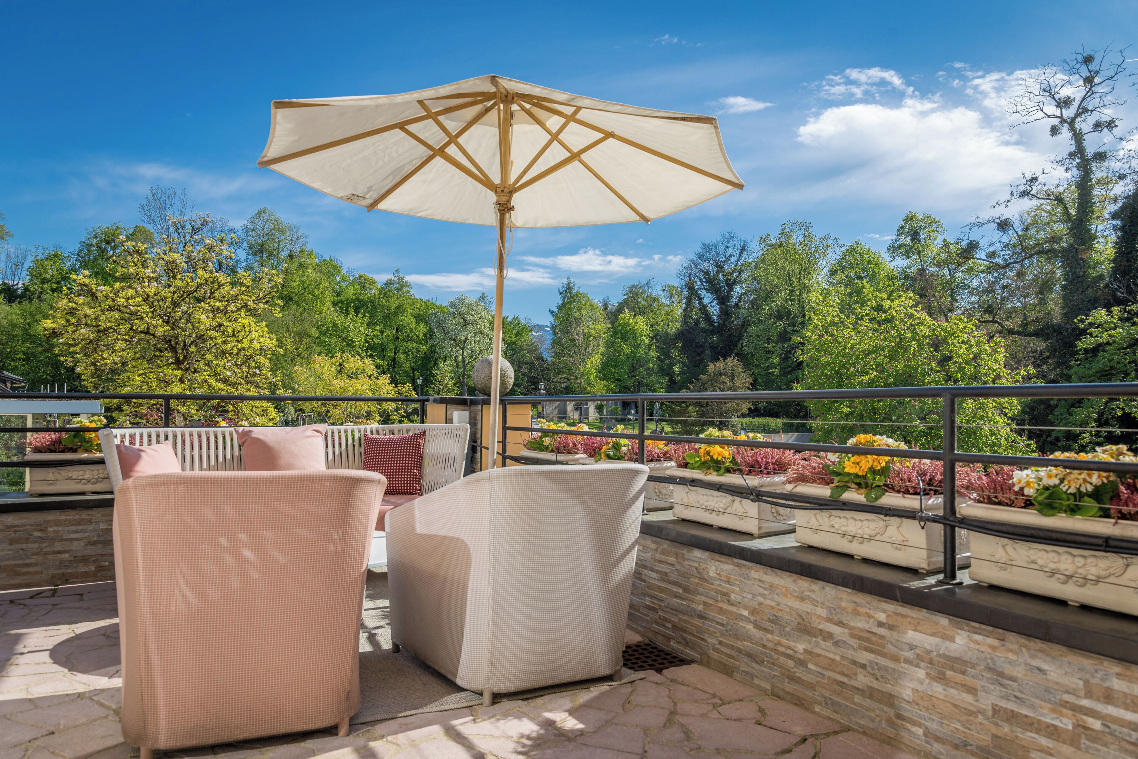 An outdoor seating area with wicker chairs and a large umbrella, overlooking a garden with various plants and trees.