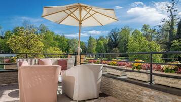 An outdoor seating area with wicker chairs and a large umbrella, overlooking a garden with various plants and trees.