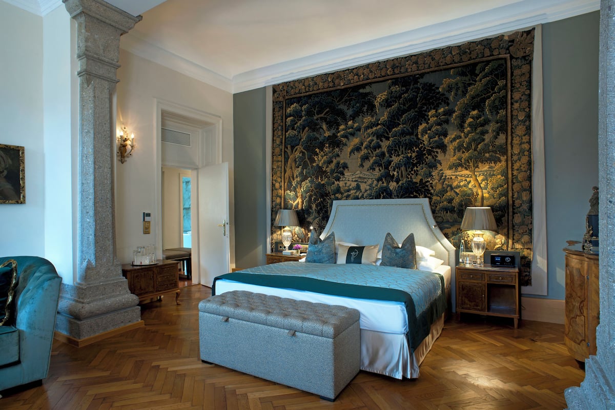 A spacious bedroom with a large bed, a nightstand, a bench, and a wall with a detailed tapestry.
