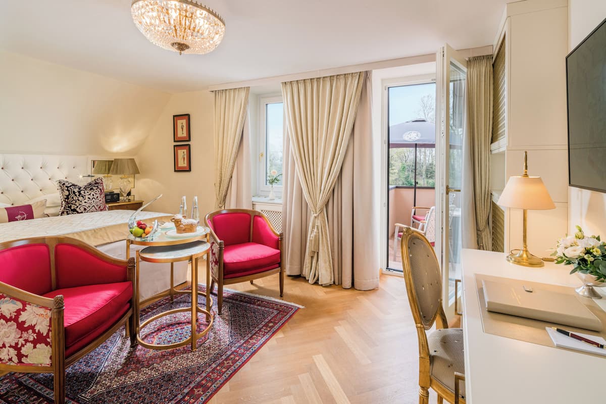 A hotel room with a large bed, a desk, a chair, a red sofa, a small table with a lamp, and a chandelier.