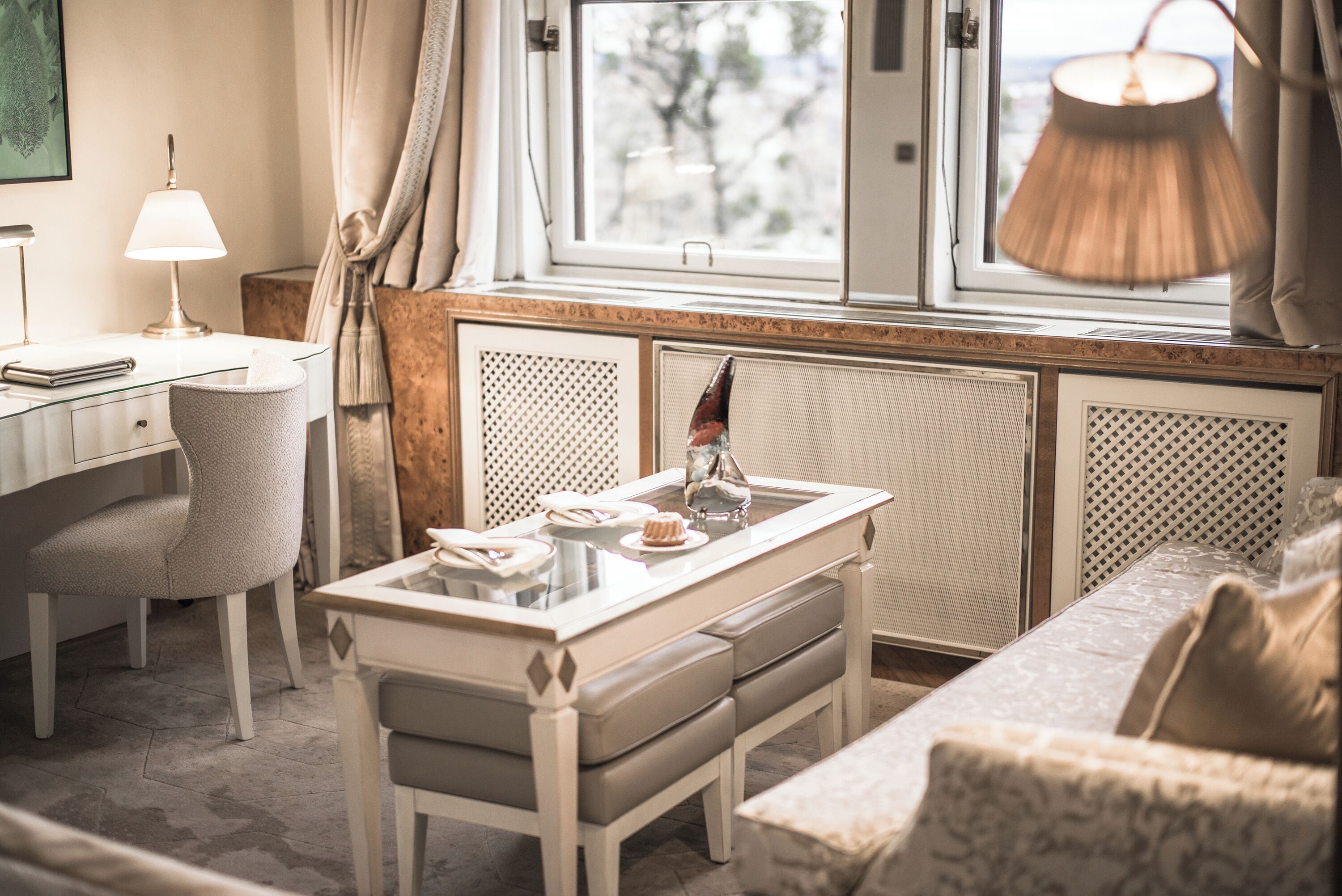 A hotel room with a dining table set for two, a chair, a sofa, a lamp, and a view of the outdoors through the window.