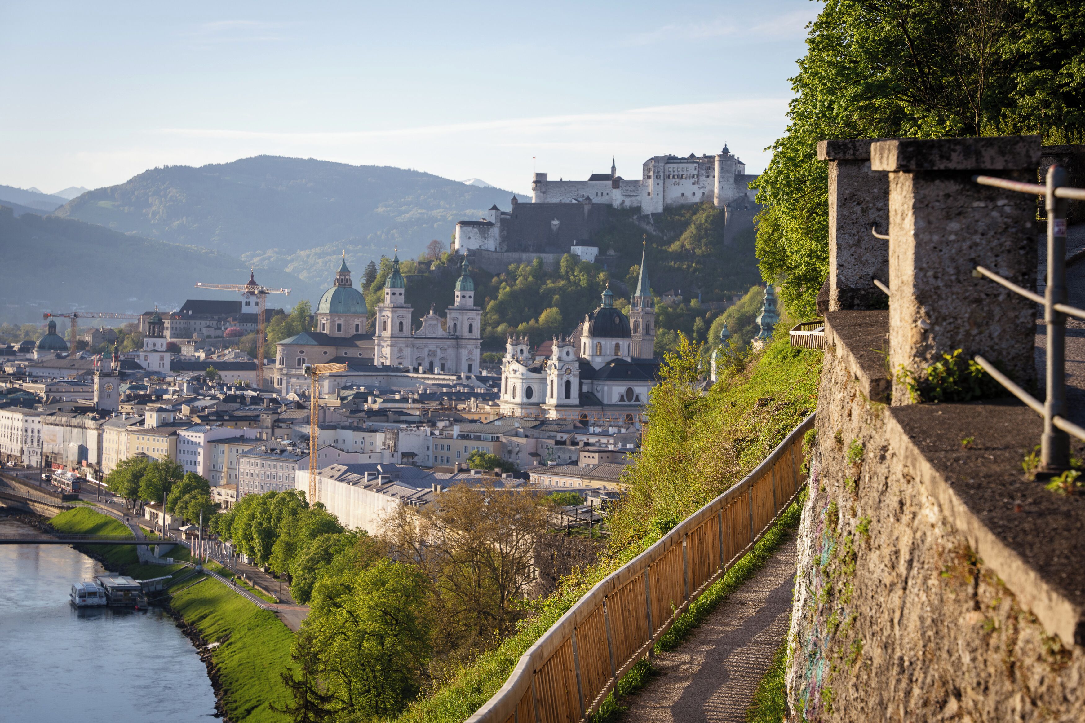 Un paisaje urbano histórico con un castillo imponente en una colina, un río y un paseo de madera a lo largo de un muro de piedra.