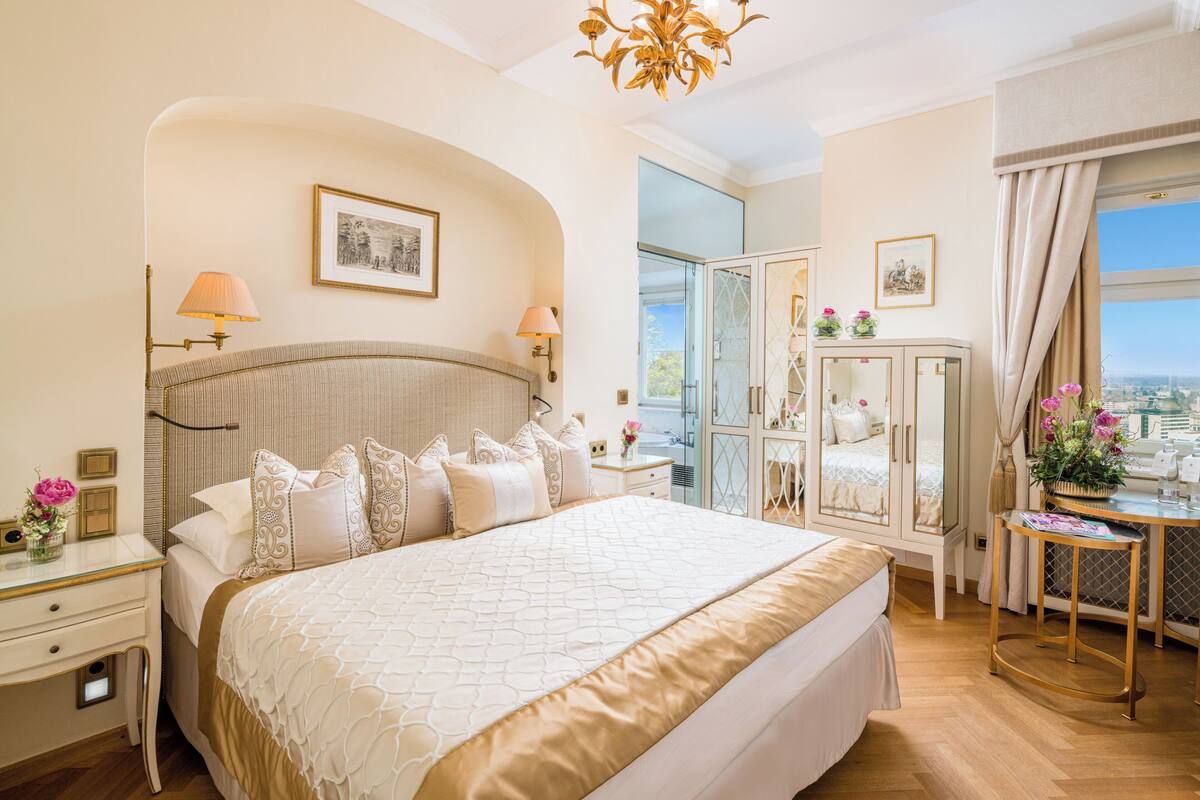 A spacious bedroom with a large bed, a vanity with a mirror, and a view of the city.