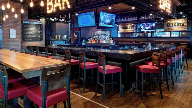 A bar area with a long wooden counter, multiple bar stools, and two televisions mounted on the wall. - Courtyard By Marriott Detroit Downtown (Detroit)