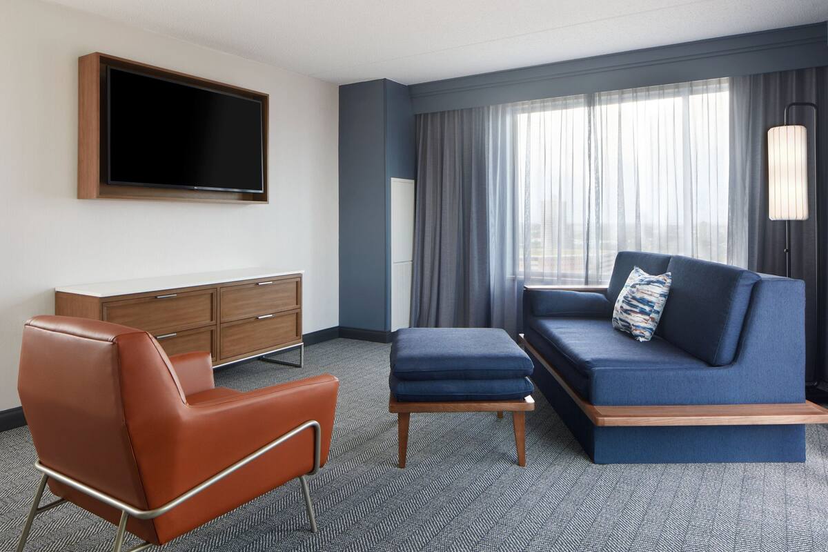 A modern hotel room with a sofa, a chair, a television, and a large window with curtains.