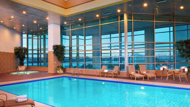 An indoor swimming pool with clear glass walls, lounge chairs, and a view of the cityscape.