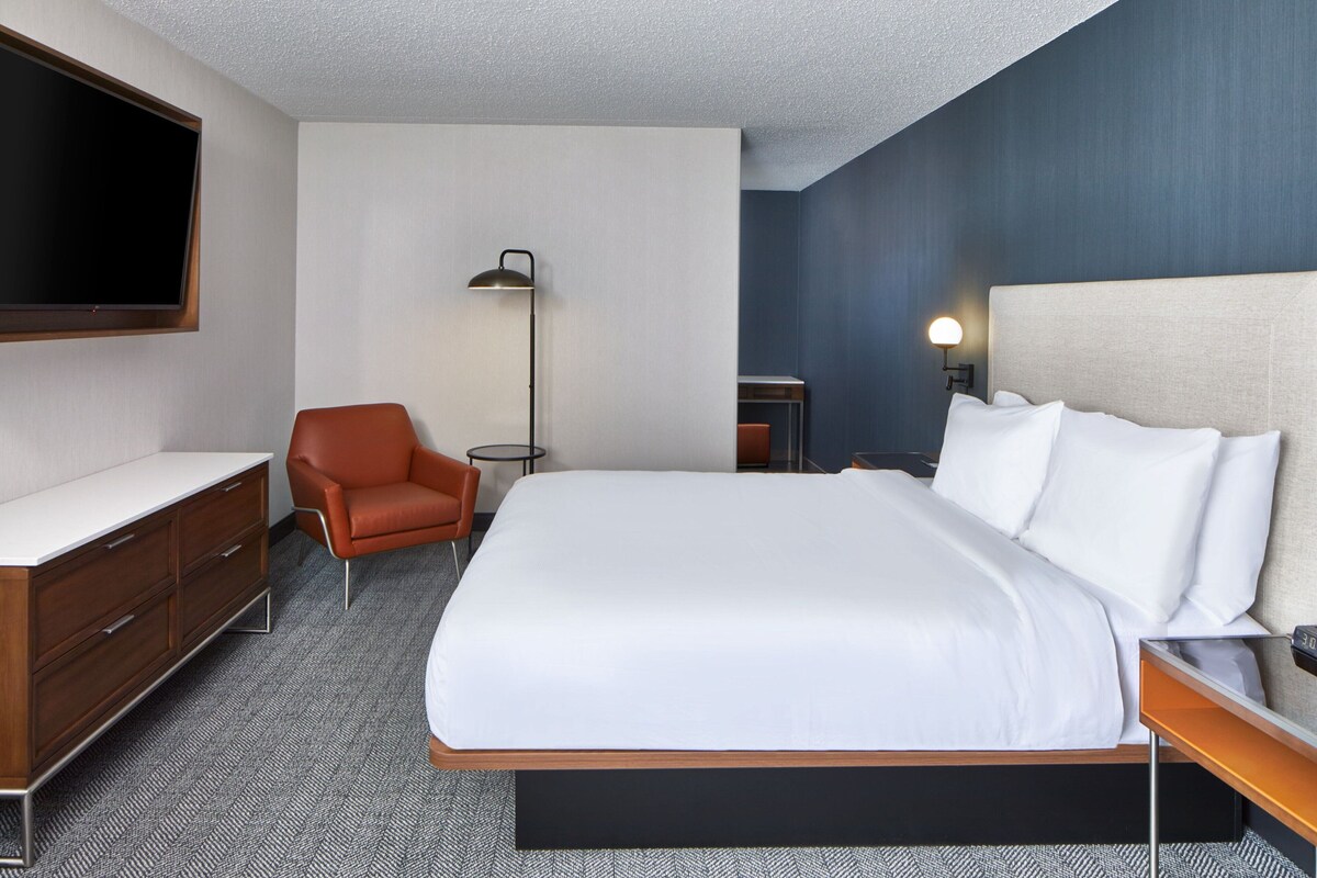 A hotel room with a large bed, a desk, a chair, a TV, and a lamp.