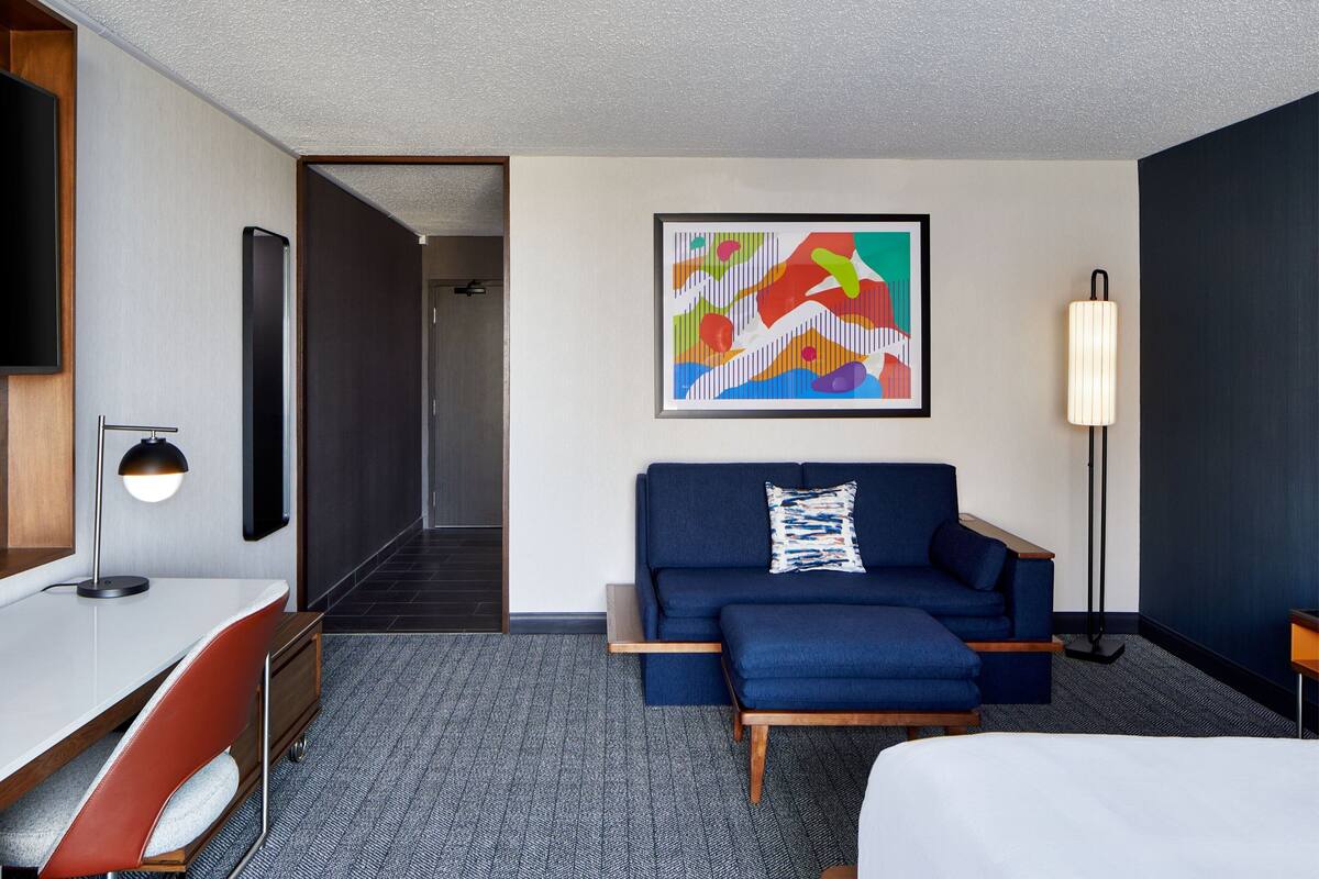 A hotel room with a bed, a desk with a chair, a sofa, a standing lamp, and a colorful abstract painting on the wall.