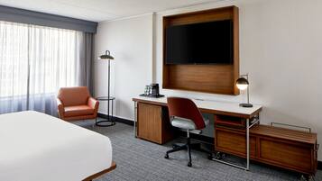 Et hotelværelse med en stor seng, et skrivebord med en computer, en stol, et fjernsyn monteret på væggen og en gulvlampe.