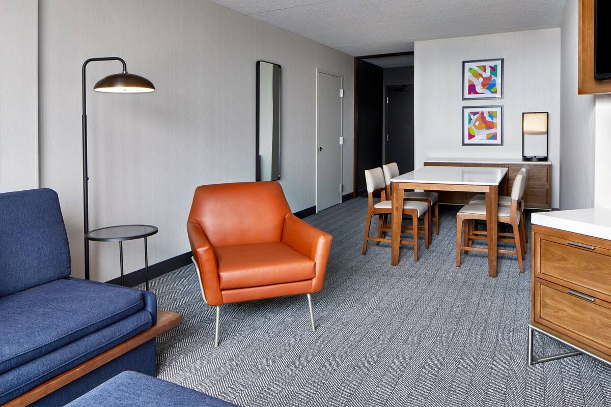 A hotel room with a blue sofa, an orange armchair, a dining table with chairs, a floor lamp, and a flat-screen TV mounted on the wall.
