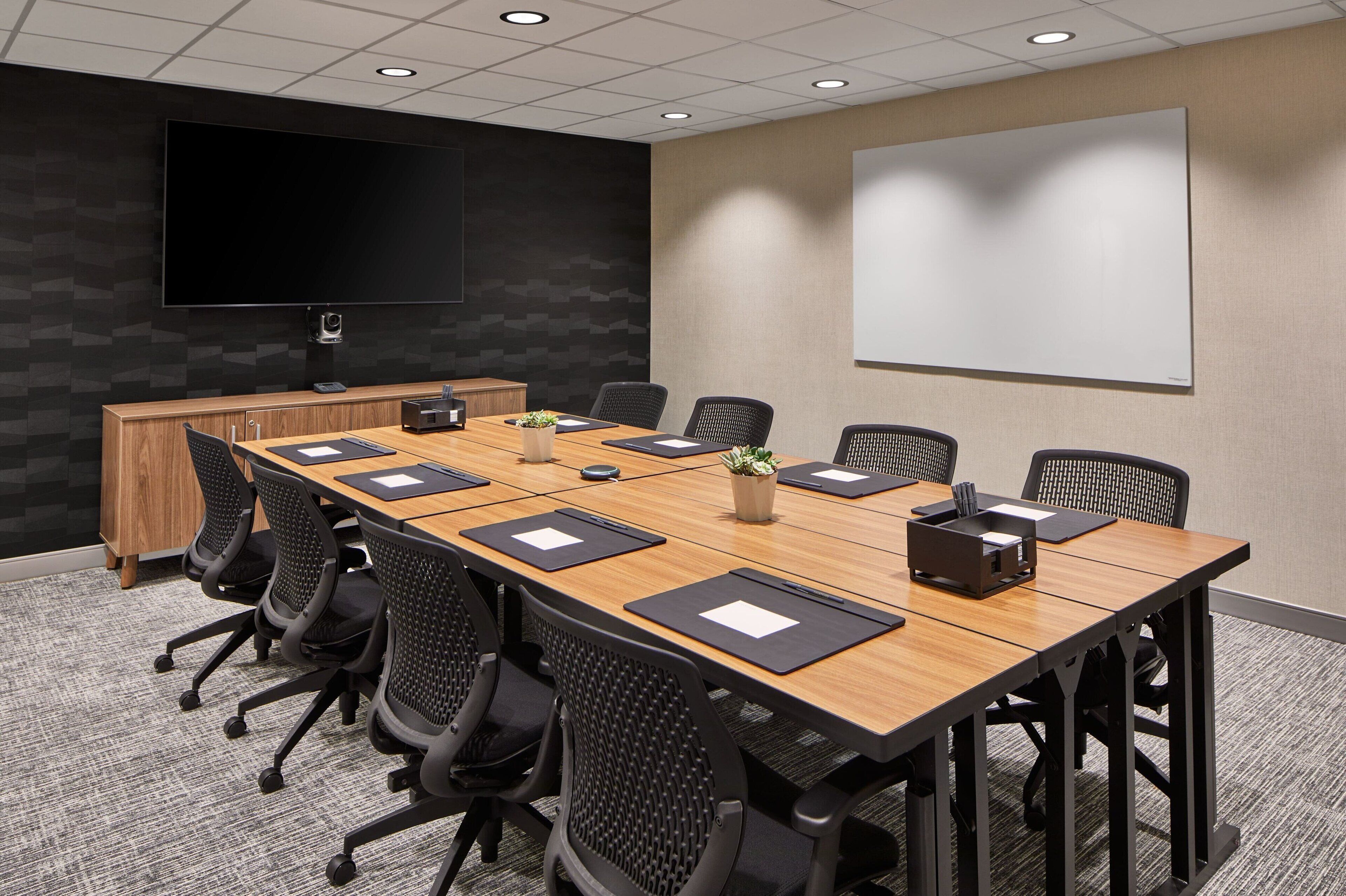 Une salle de conférence avec une longue table, des chaises noires, un bureau en bois, un téléviseur à écran plat et un tableau blanc.