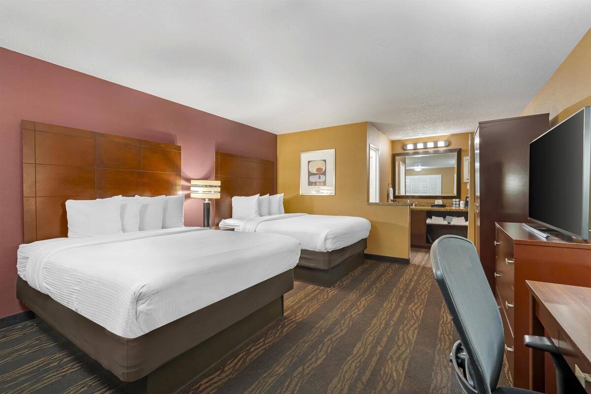 A hotel room with two beds, a desk, a chair, a TV, a mirror, and a bathroom with a toilet and sink.