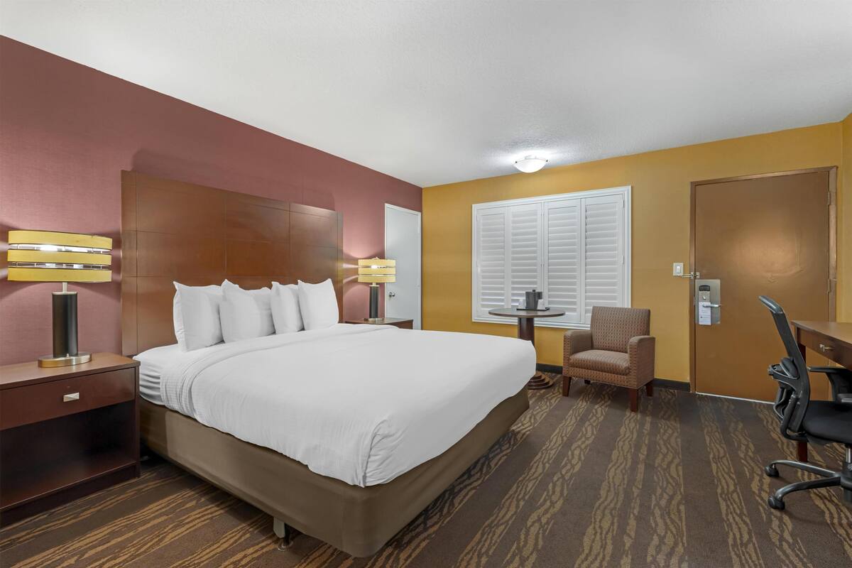 A hotel room with a large bed, a desk, a chair, a small table, and a door.