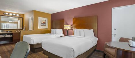 A hotel room with two beds, a desk, a chair, a small table, a mirror, and a lamp.