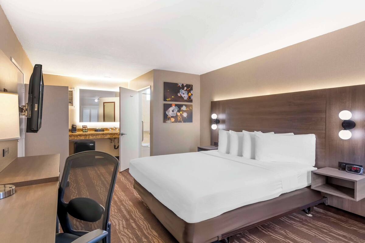 A hotel room with a large bed, a desk with a chair, a TV, and a bathroom visible through an open door.